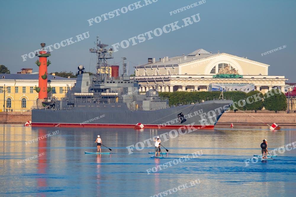 На фотографии изображен военный корабль, стоящий на реке Нева у Стрелки Васильевского острова в Санкт-Петербурге. На переднем плане видны люди, катающиеся на SUP-досках. Виден один из Ростральных колонн и здание Биржи на заднем плане.